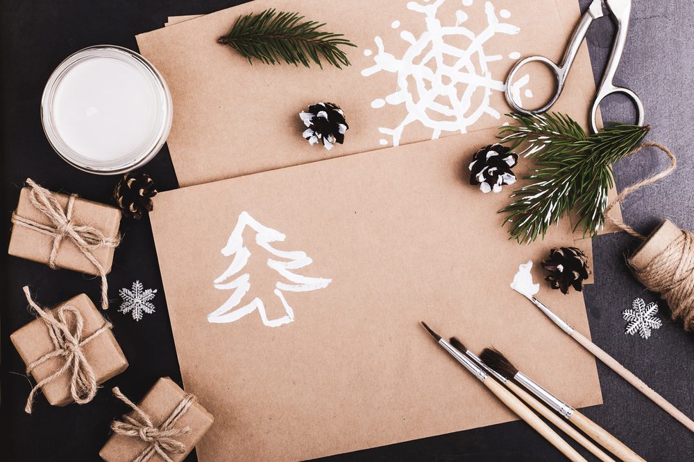 Craft supplies arranged neatly on a table; brown paper features white paint snowflakes and tree, with brushes, twine, small pine cones, and greenery.