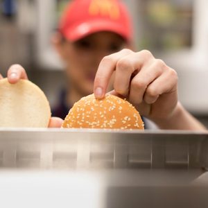 Hands place burger buns on a tray; a person wears a red cap in a kitchen setting.