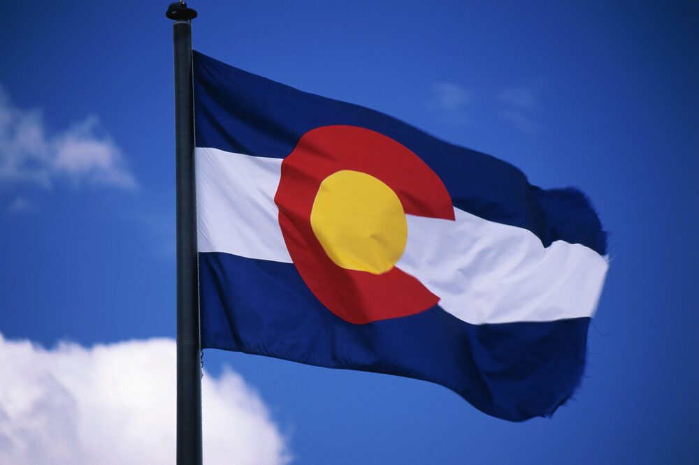 This is the Colorado State Flag, waving in the wind situated on a flag pole. It is set against a blue sky. At the center of the flag is a large capital C.