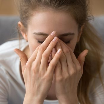 A person covers their face with hands, expressing stress, in a neutral indoor setting.