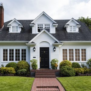 A white house with a black door and chimney sits amidst lush greenery, featuring a brick path and manicured lawn.