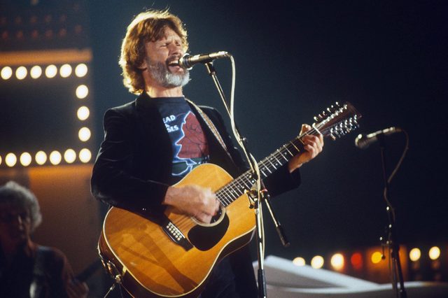 Musician sings passionately while playing guitar on stage, surrounded by bright lights.