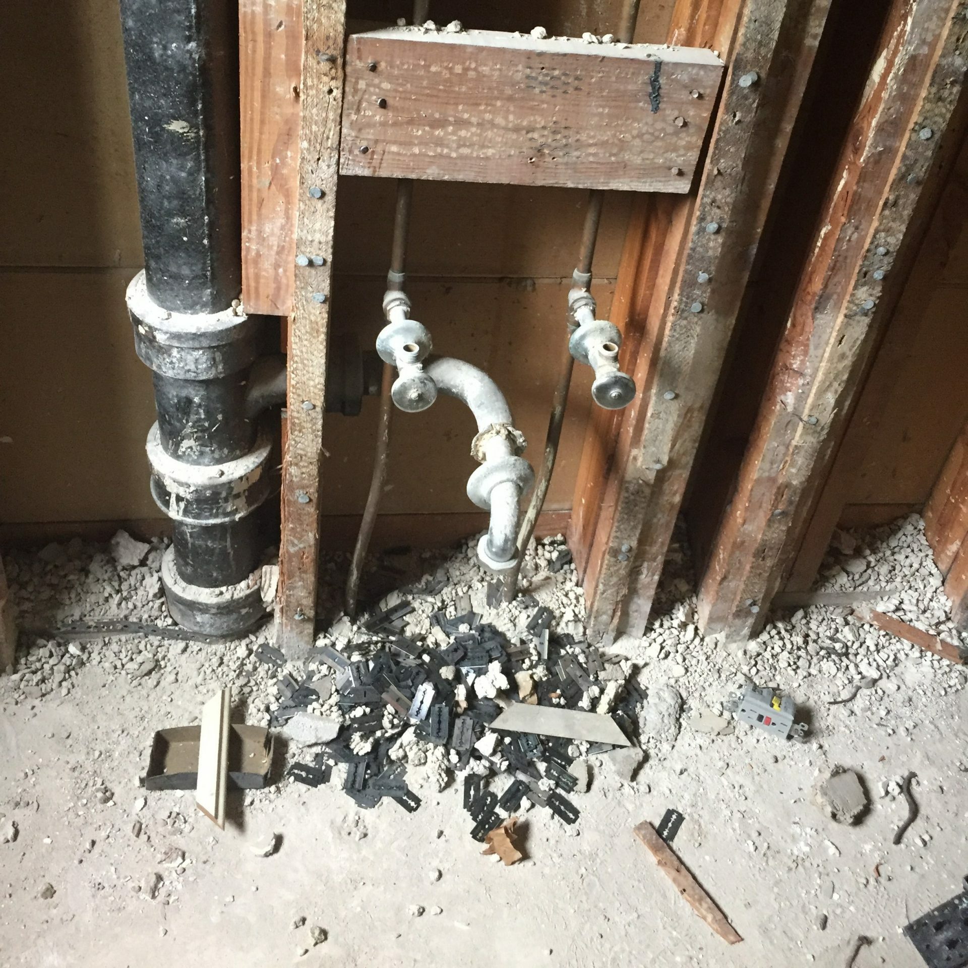 Exposed water pipes protrude from a wooden wall structure, surrounded by debris and broken black plastic pieces on a dusty floor.
