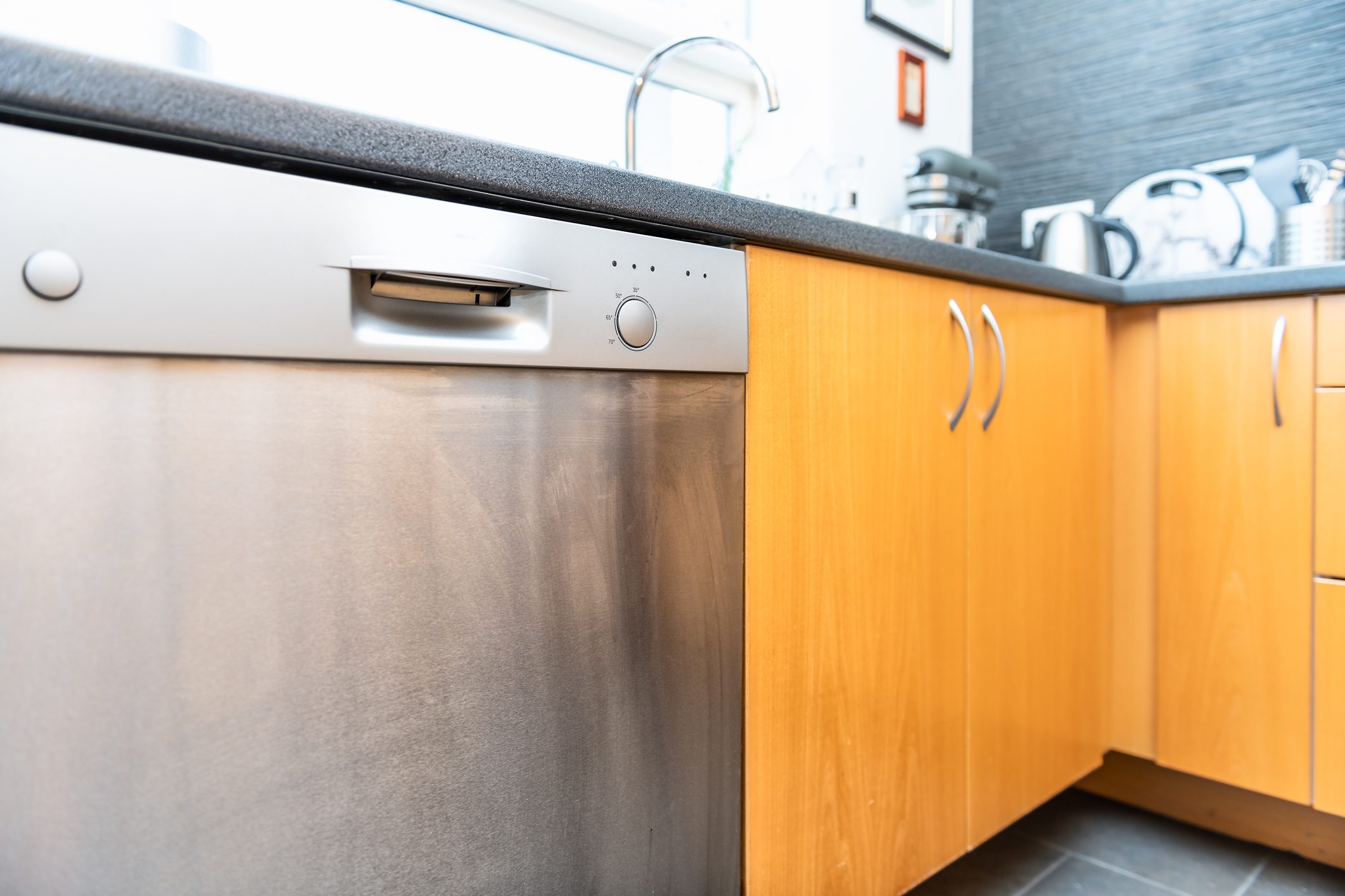 Closeup of side of modern wood, wooden finish kitchen drawers with steel handles, cabinets, dishwasher, tile, tiled brown, yellow floor, utensils, faucet