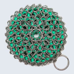 Metal chainmail scrubber rests flat, with a circular pattern on a teal surface. A small ring attached displays text.