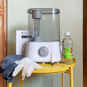 Humidifier sits on a yellow table, surrounded by a cleaning cloth, gloves, vinegar bottle, and paper, against a wooden door background.