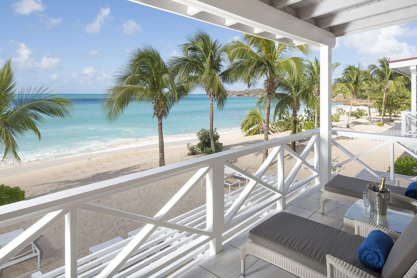 Chairs rest on a beachfront balcony, overlooking palm trees and turquoise ocean under a clear sky, with a champagne bucket and glasses nearby.