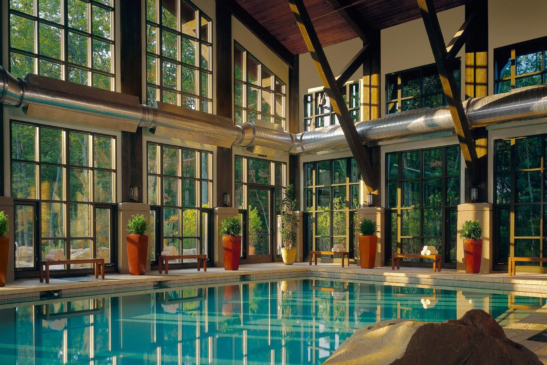 Indoor pool reflects sunlight; surrounded by tall windows, benches with towels, and potted plants; natural light fills the spacious, wood-accented room.