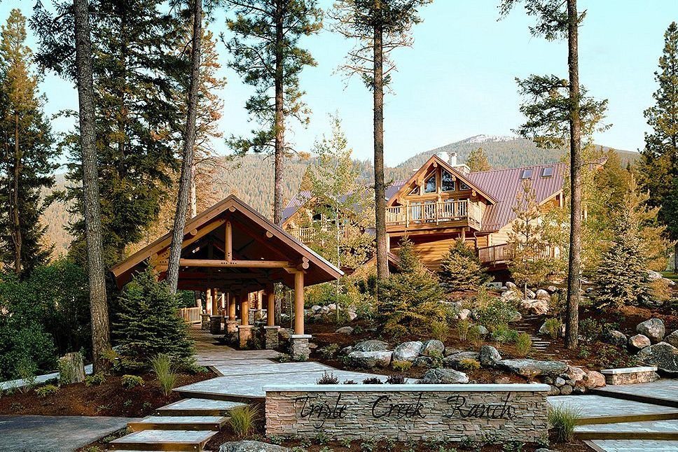 Lodge surrounded by pine trees, rests on a mountainous landscape. Stone path leads to entry, with text "Triple Creek Ranch" on a stone wall.