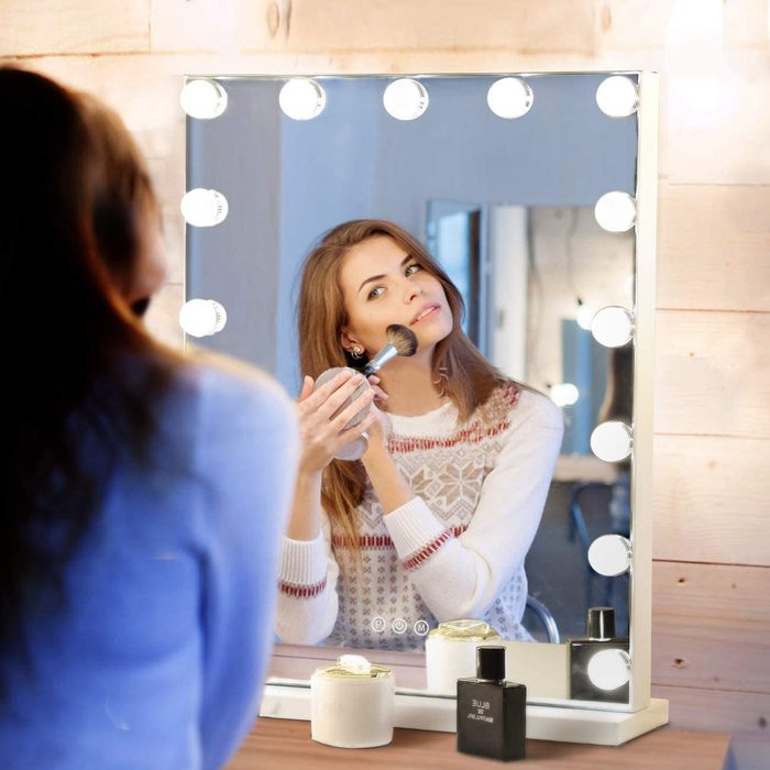 Woman applies makeup using brush, facing illuminated mirror, in a wooden-walled room. Perfume and jar are visible on the dressing table.