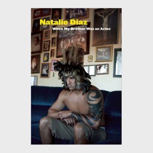 Shirtless man with tribal tattoo, wearing feathered headdress, sits on couch in front of wall covered with framed photos. Text reads 