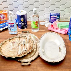 Cleaning supplies and utensils are arranged on a wooden countertop against a white tiled wall, including baking soda, salt, vinegar, and cleaning liquids.