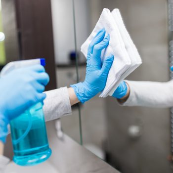 Gloved hands clean a mirror with a cloth and spray bottle in a bathroom setting.