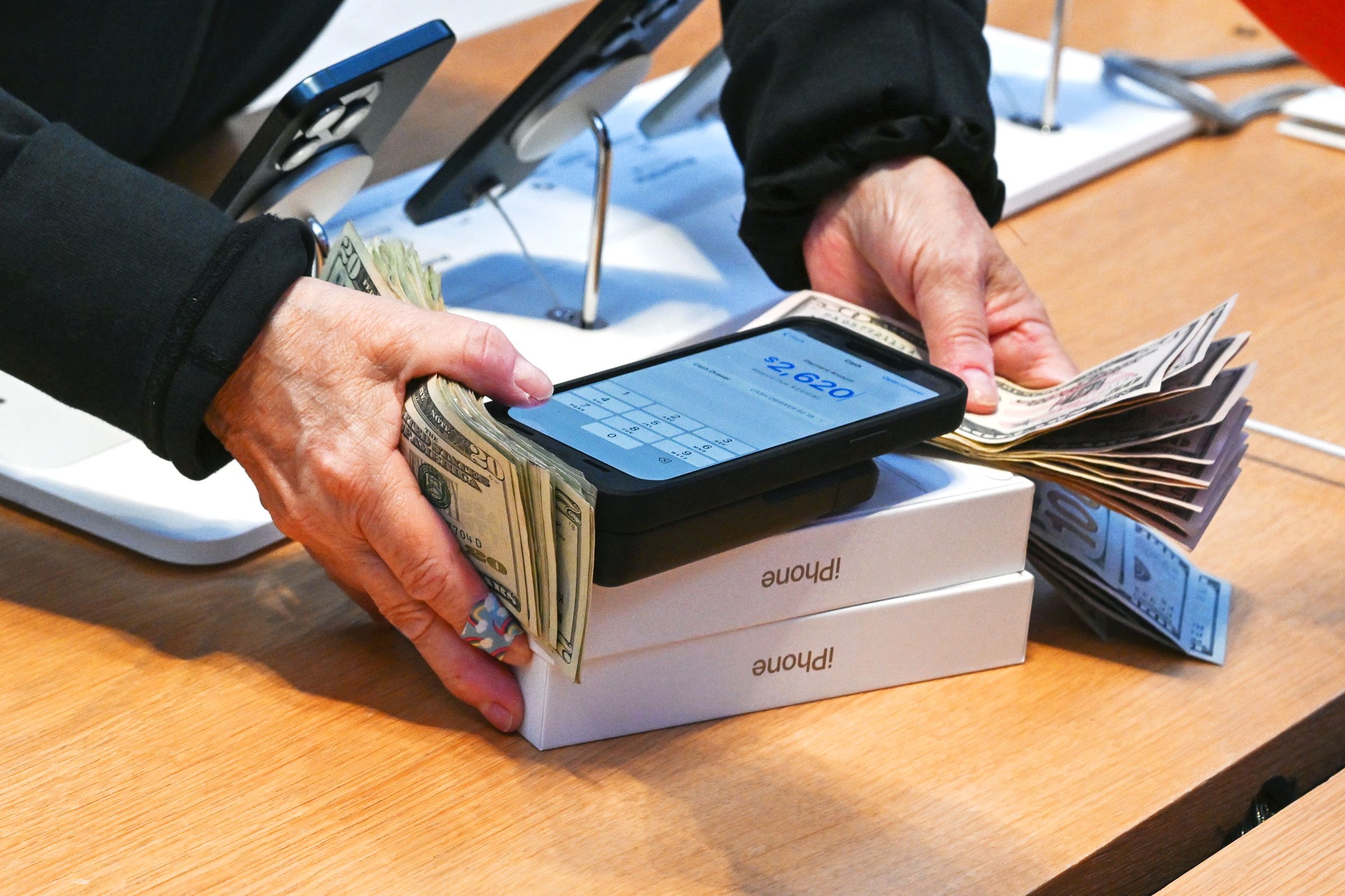 Hands holding cash and a smartphone showing $2,620; on iPhone boxes, surrounded by electronic devices on a store counter.