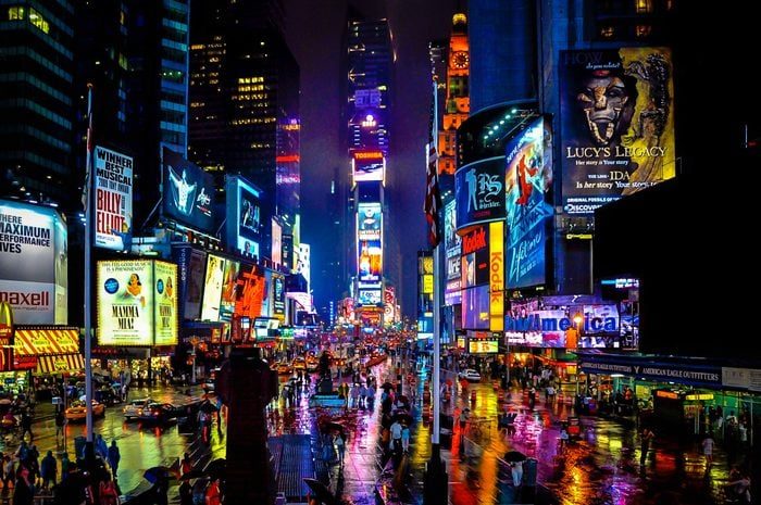 Neon billboards illuminate bustling crowds on a wet street in Times Square at night, creating vibrant reflections on the pavement.