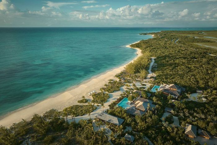 Beach stretches along turquoise ocean; lush greenery surrounds villas and pools, creating a serene coastal retreat under partly cloudy skies.