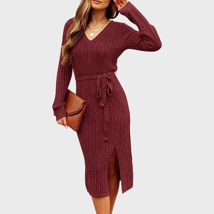 A woman poses wearing a burgundy cable-knit dress with a belt and slit, holding a brown clutch, against a plain background.