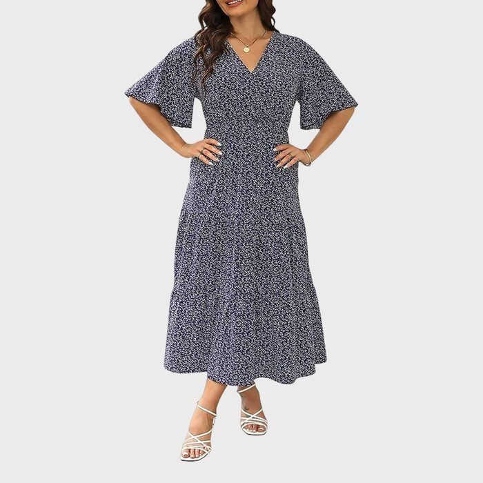 A woman models a navy floral dress with short sleeves and a V-neck, standing against a plain background, wearing white strappy sandals.