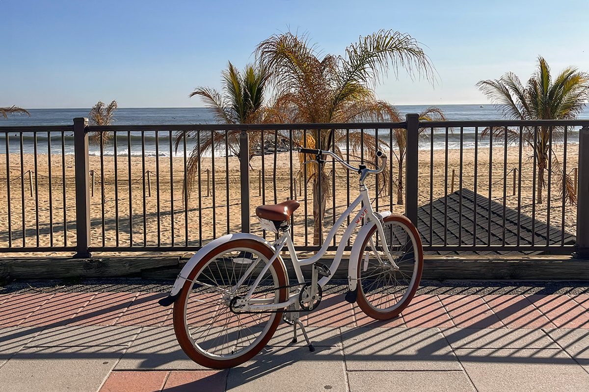 A white bicycle with brown accents is parked on a paved walkway by a metal railing. Behind the railing, there is a sandy beach with several palm trees and a calm ocean under a clear blue sky.