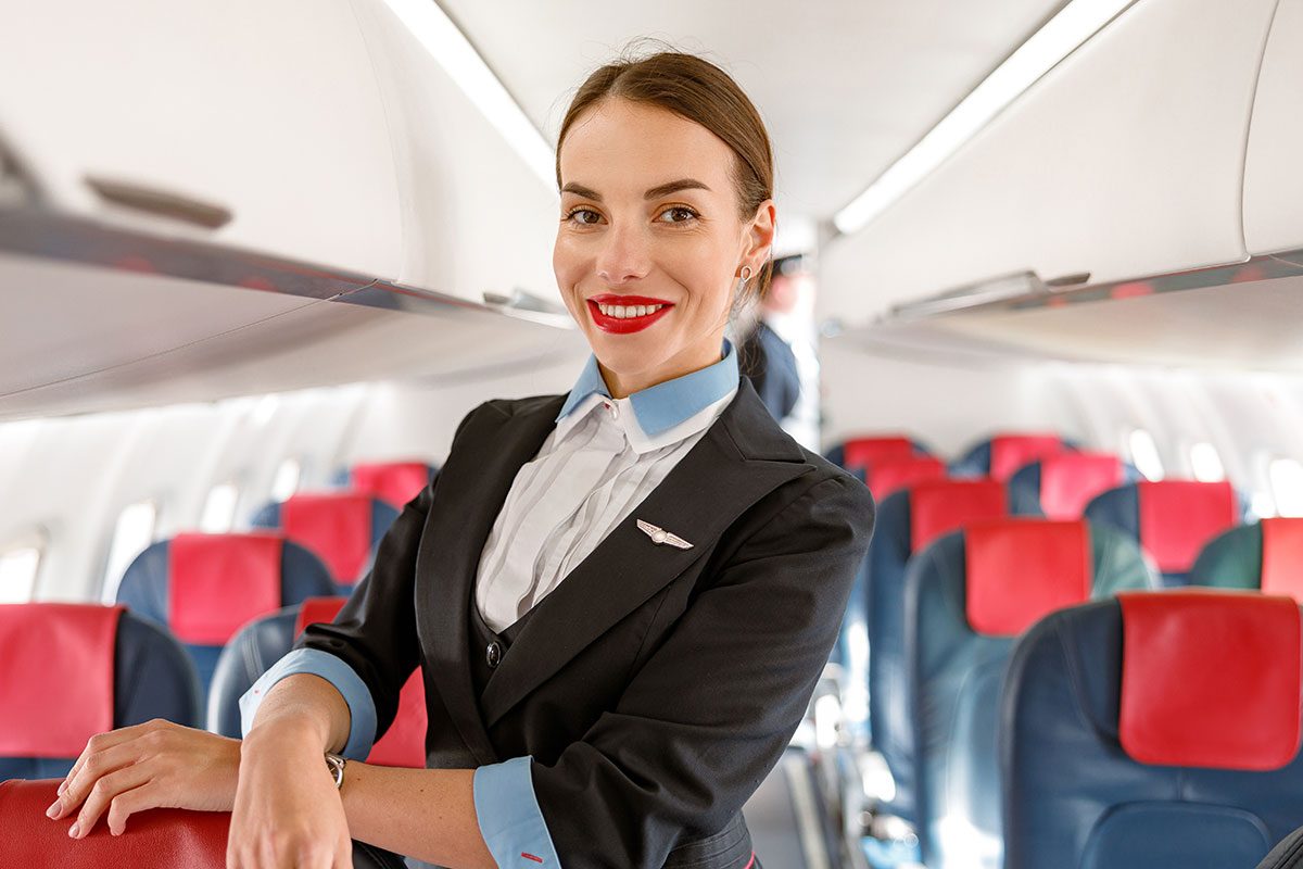 Cheerful woman stewardess or air hostess looking at camera and smiling while placing hand on passenger chair in aircraft